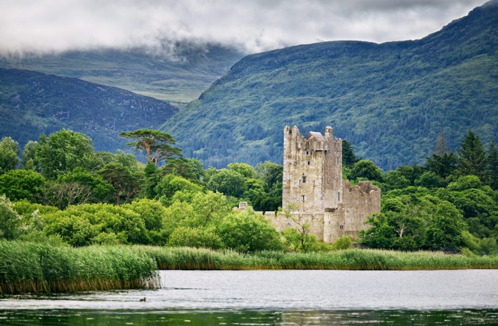 A stone castle tower surrounded by green trees beside a lake with mountains